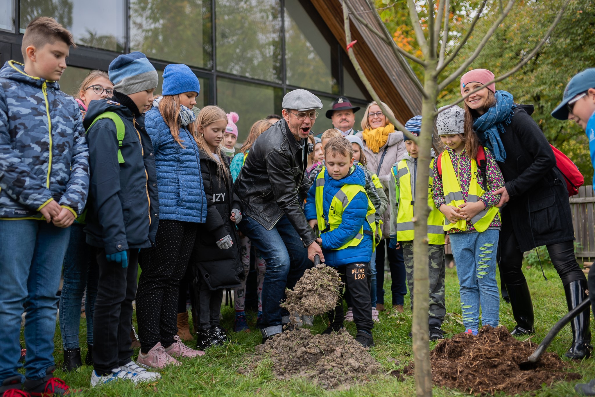 Zdjęcie przedstawia grupę osób w różnym wieku sadzących drzewo, w tle widać budynek SDK.