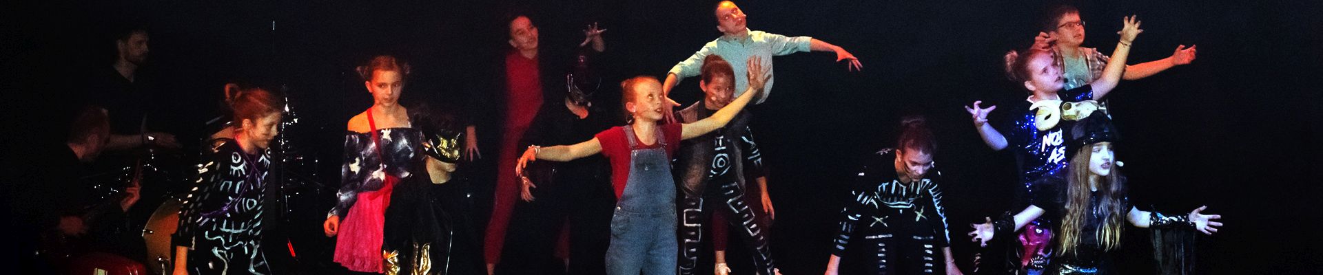 Na zdjęciu członkowie grupy musicalowej na scenie odgrywają dynamiczny układ choreograficzy, z rękami podniesionymi ku górze. Ciemne tło.