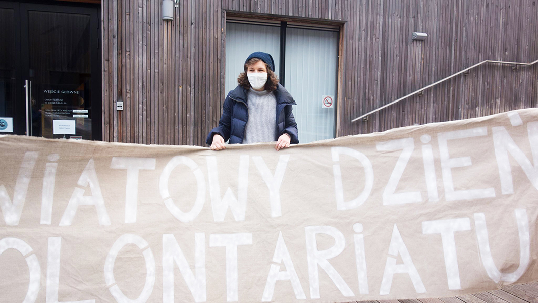 Finał projektu „JESTEŚMY TU!”; na zdjęciu wolontariuszka z wielkim papierowym transparentem z napisem "Światowy Dzień Wolontariatu".Foto Dominika Dzieniszewska.