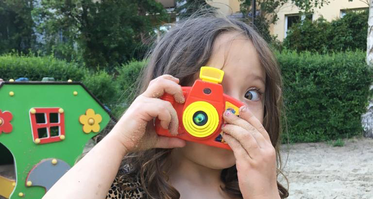 Na zdjęciu fragmeng twarzy dzieczynki trzymającej aparat fotograficzny przy twarzy.