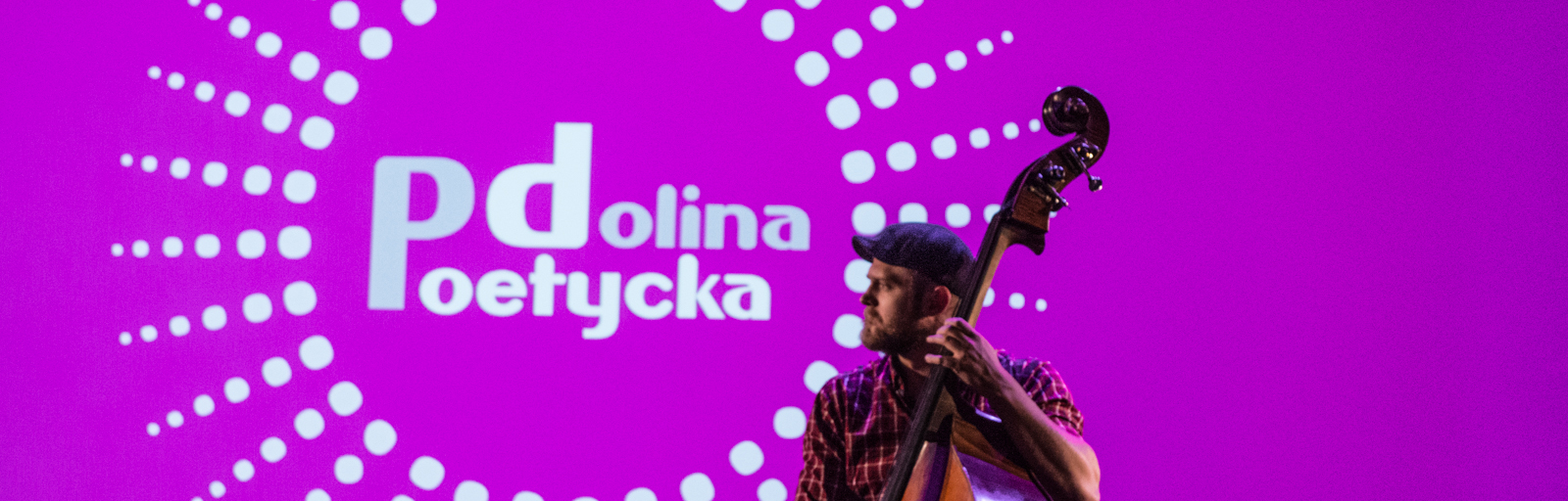 Zdjęcie przedstawia młodego mężczyznę grającego na wiolonczeli.. W tle różowa plansza, z logotypem Festiwalu Poetycka Dolina,  ksztłcie słońca, z napisem w centralnym miejscu. 