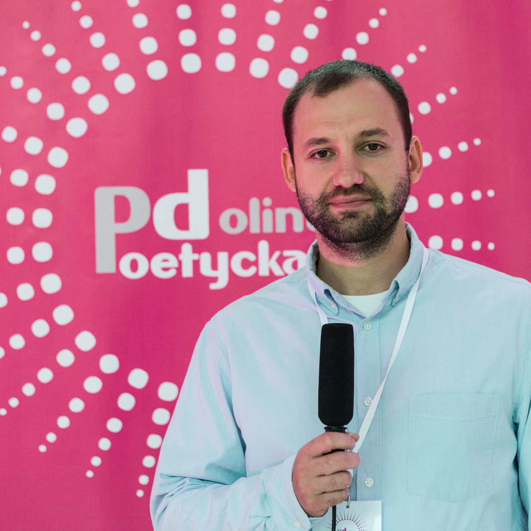 Zdjęcie przedstawia młodego mężczyznę z mikrofonem, występującego w roliw roli konferansjera. W tle różowa plansza, z logotypem Festiwalu Poetycka Dolina,  ksztłcie słońca, z napisem w centralnym miejscu. 