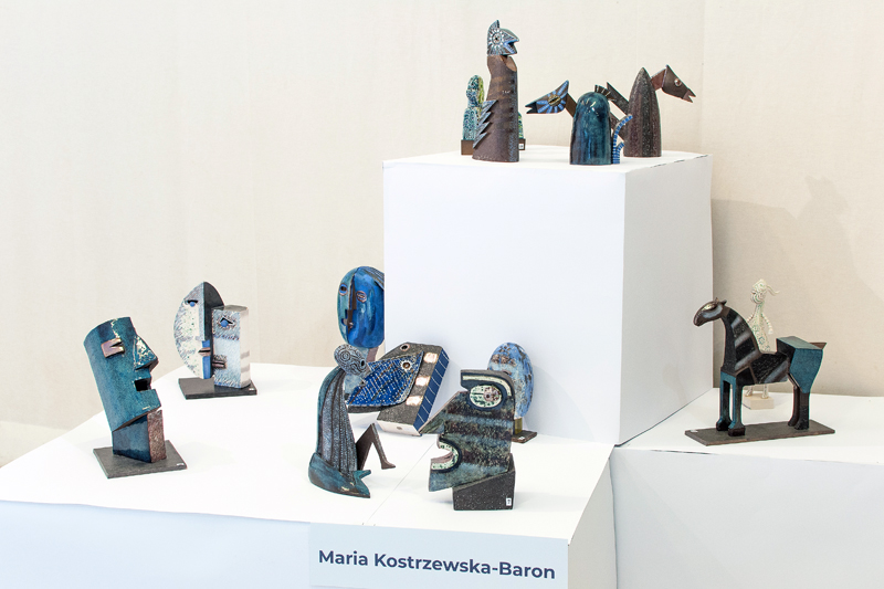 Zdjęcie przedstawiające ekspozycję ceramicznych rzeźb Marii Kostrzewskiej-Baron. Prace wyeksponowano na dwóch poziomach: górny to artystycznie przetworzone wizerunki ptaków; w dolnej cześci widzimy ceramiczne figuracje portretowe, siedzącą postać ludzką oraz – z lewej strony – ceramiczną rzeźbę konia i w tyle jasną figurkę kobiecą.