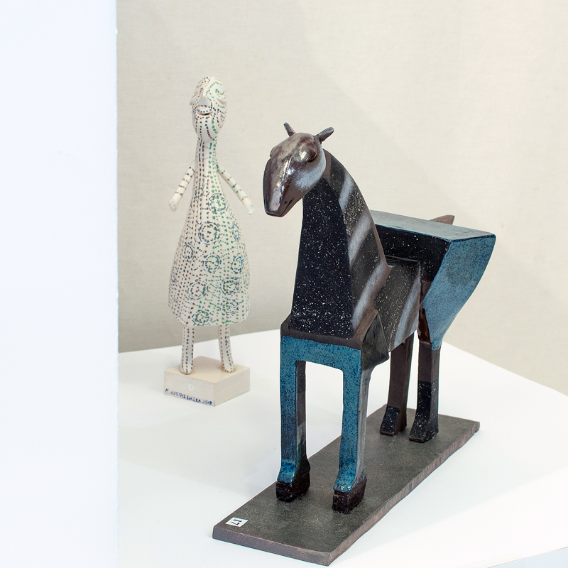 Ceramiczna rzeźba konia utrzymana w kolorach brązu i błękitu. Obok figurka kobieca w bieli. Powierzchnia rzeźby malowana w delikatne kwiatowe wzory.
