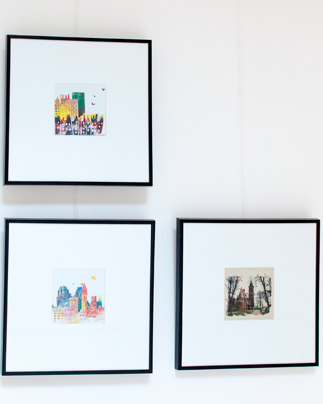 Zdjęcie przedstawiające ekspozycję trzech obrazów. Z lewej strony, jeden pod drugim, prezentowane są dwa syntetyczne w ujęciu pejzaże wielkomiejskie z grupami wysokich, kolorowych wieżowców. Trzeci obraz to także przedstawienie architektury. Widnieje na nim wiejski kościółek w otoczeniu bezlistnych drzew, widziany od strony absydy. Stonowany w gamie barwnej obrazek, utrzymany w tonach brunatnych i sepii.