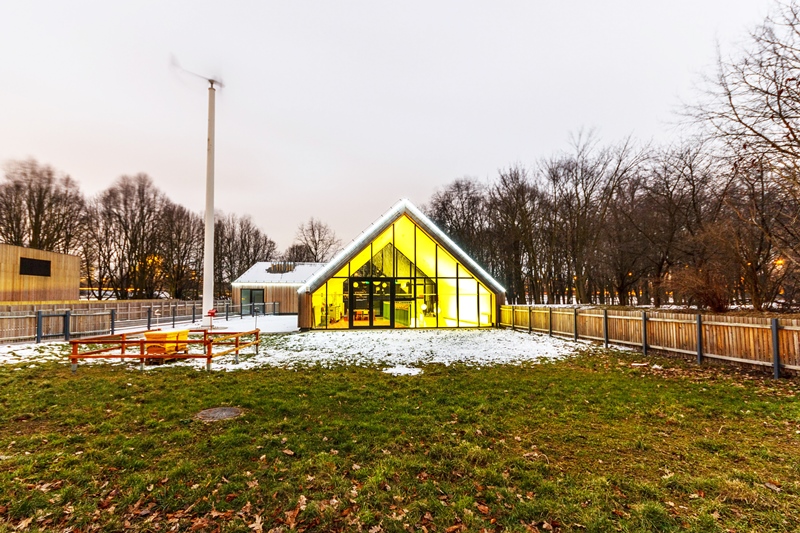 Zdjęcie siedziby SDK Na zdjęciu budynek edukacyjny widziany z daleka. Obok elektrownia wiatrowa oraz ule dla pszczół.