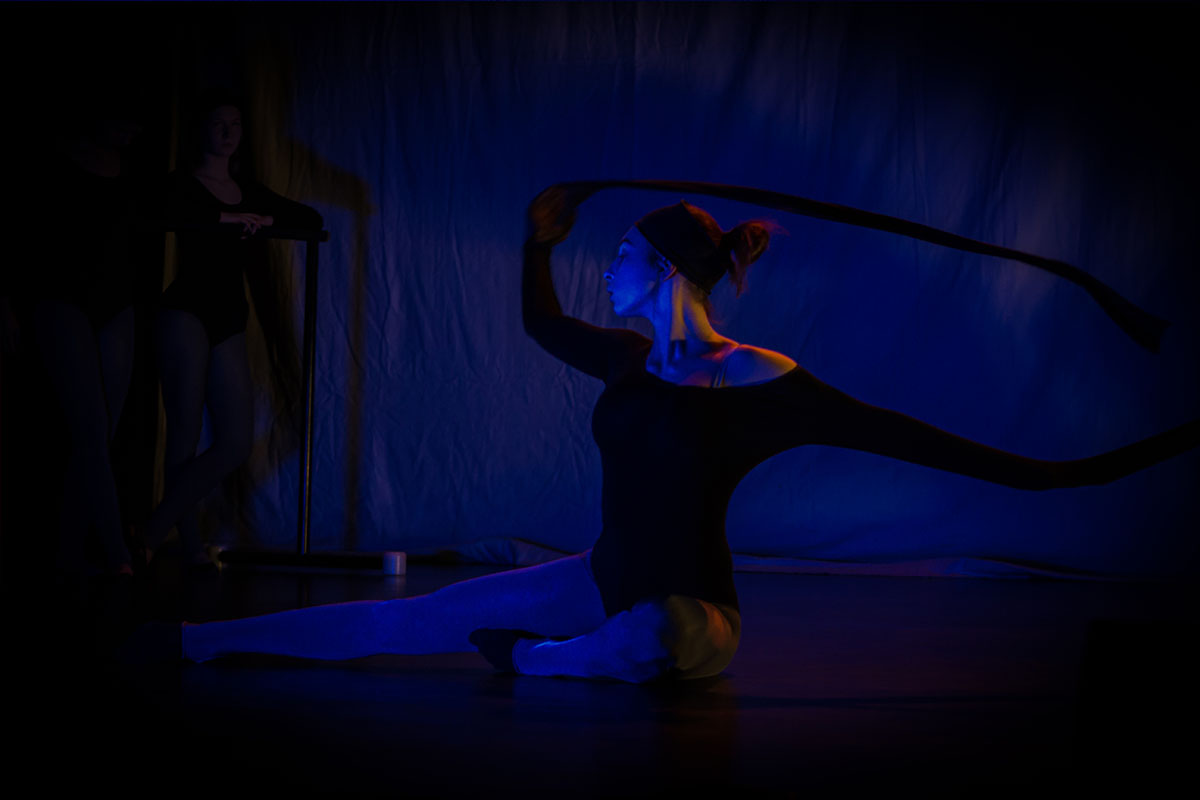Na zdjęciu widoczna jest tancerka siedząca na scenie z rękami podniesionymi do góry. Na scenie przeważa niebieskie, ciemne światło. 