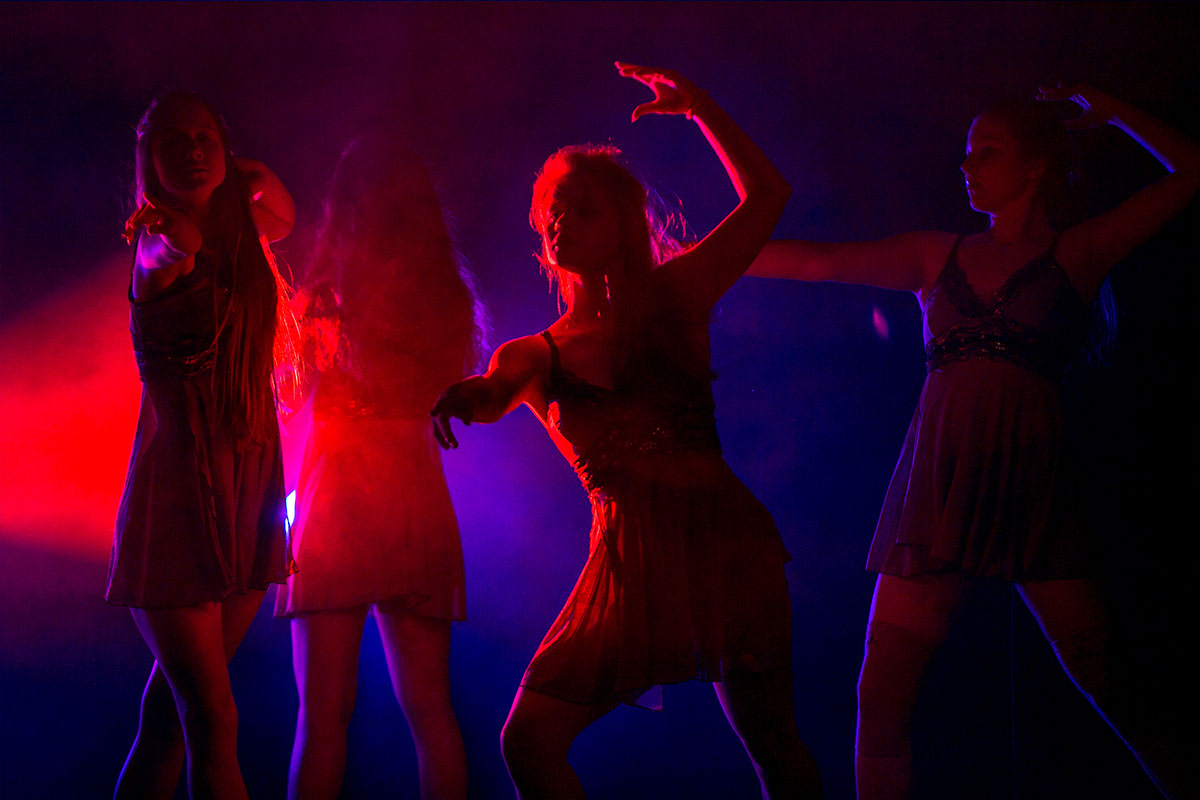 Zdjęcie przedstawia cztery dziewczyny w tanecznych pozach na scenie. Na zdjęciu przeważa czerwony kolor światła. 