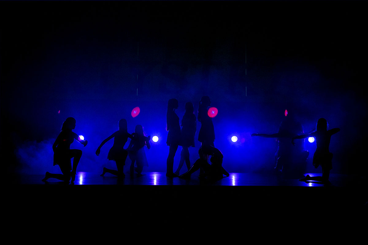 Fotografia przedstawia grupę osób na scenie. Widoczne są ich same kontury. Na scenie przeważa niebieskie światło. 