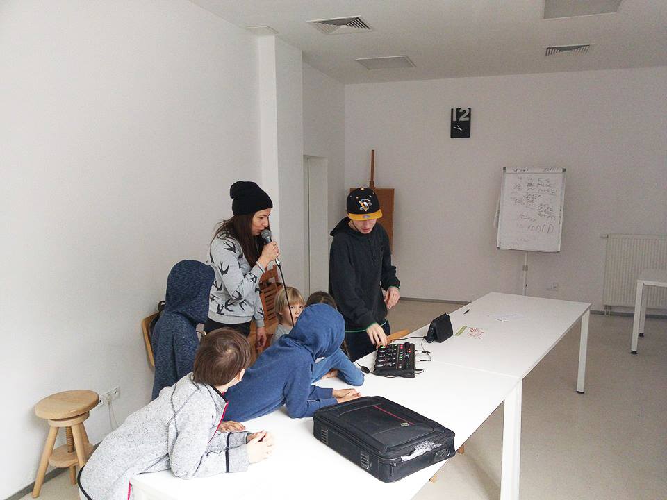 Na zdjęciu widoczna jest grupa osób stojących wokół stołu, na którym znajduje się sprzęt do beatboxu. Dziewczyna trzyma w ręku mikrofon. 