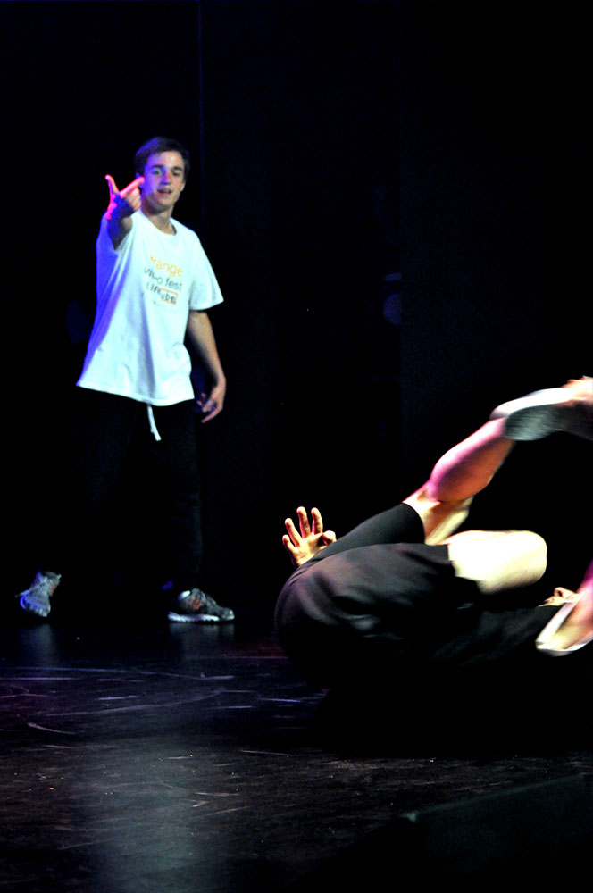 Na zdjęciu widocznych jest dwóch tancerzy. Na pierwszym planie tancerz w pozycji akrobatycznej, na dalszym planie drugi tancerz stojący w normalnej pozycji. 