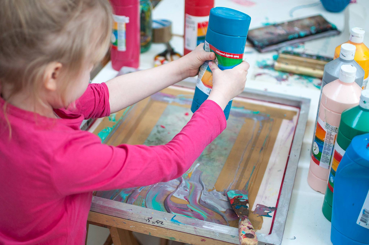 Na zdjęciu widoczna jest dziewczynka wyciskające farbę na sito od sitodruku. 