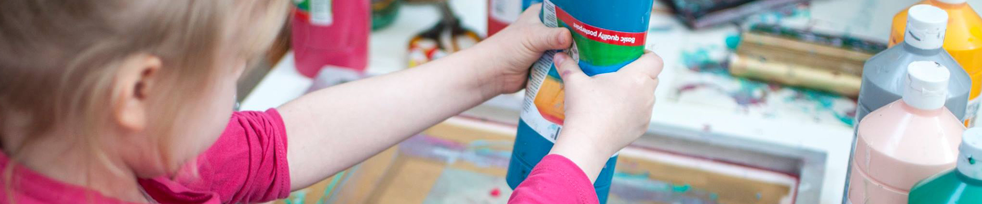 Na zdjęciu dziecko widziane od tyłu wylewające farbę na powstałą pracę plastyczną. W tle przybory do warsztatu.
