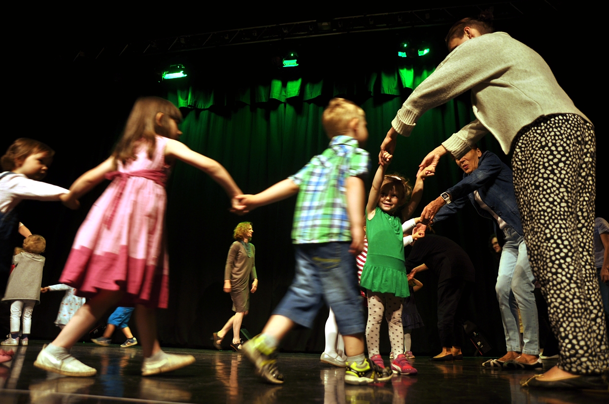 Na zdjęciu widać sylwetki dzieci tańczące w sali widowiskowej. Dzieci zostały ustawione w kółku i prowadzi je osoba dorosła.