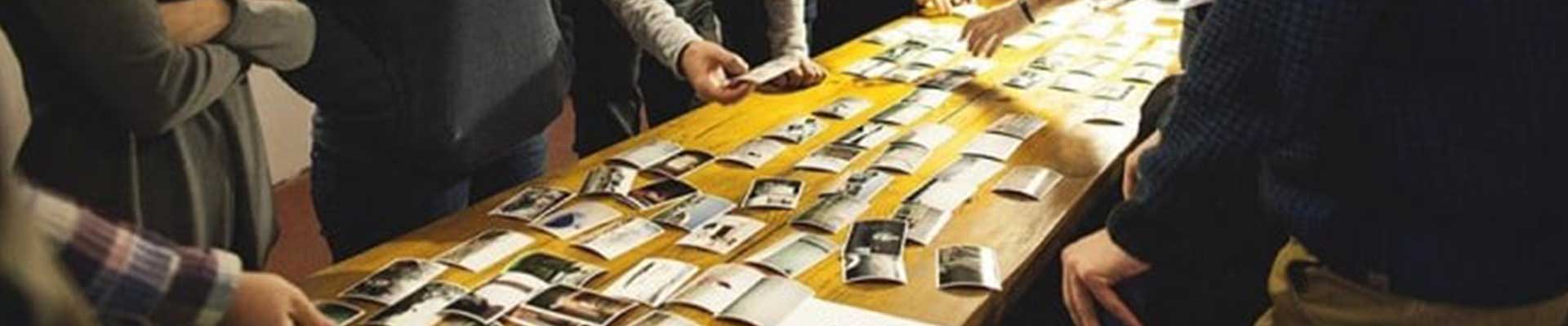 Na zdjęciu grupa osób stojąca przy stole podczas wyboru zdjęć, które ułożone są na stole. Nie widoczne są twarze uczestników.