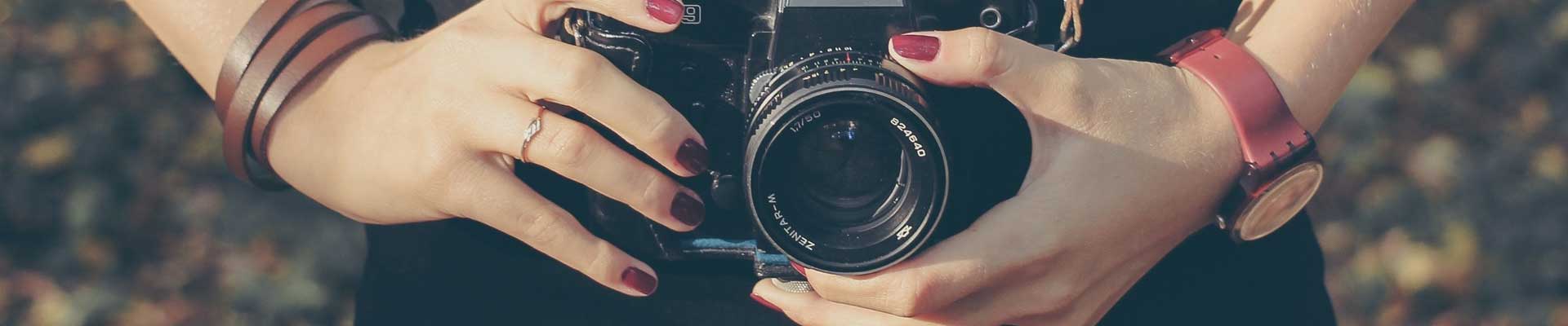 Zdjęcie: fragment kobiecej postaci trzymającej w dłoniach z pomalowanymi paznokciami aparat fotograficzny. Fot. za pixaby.pl