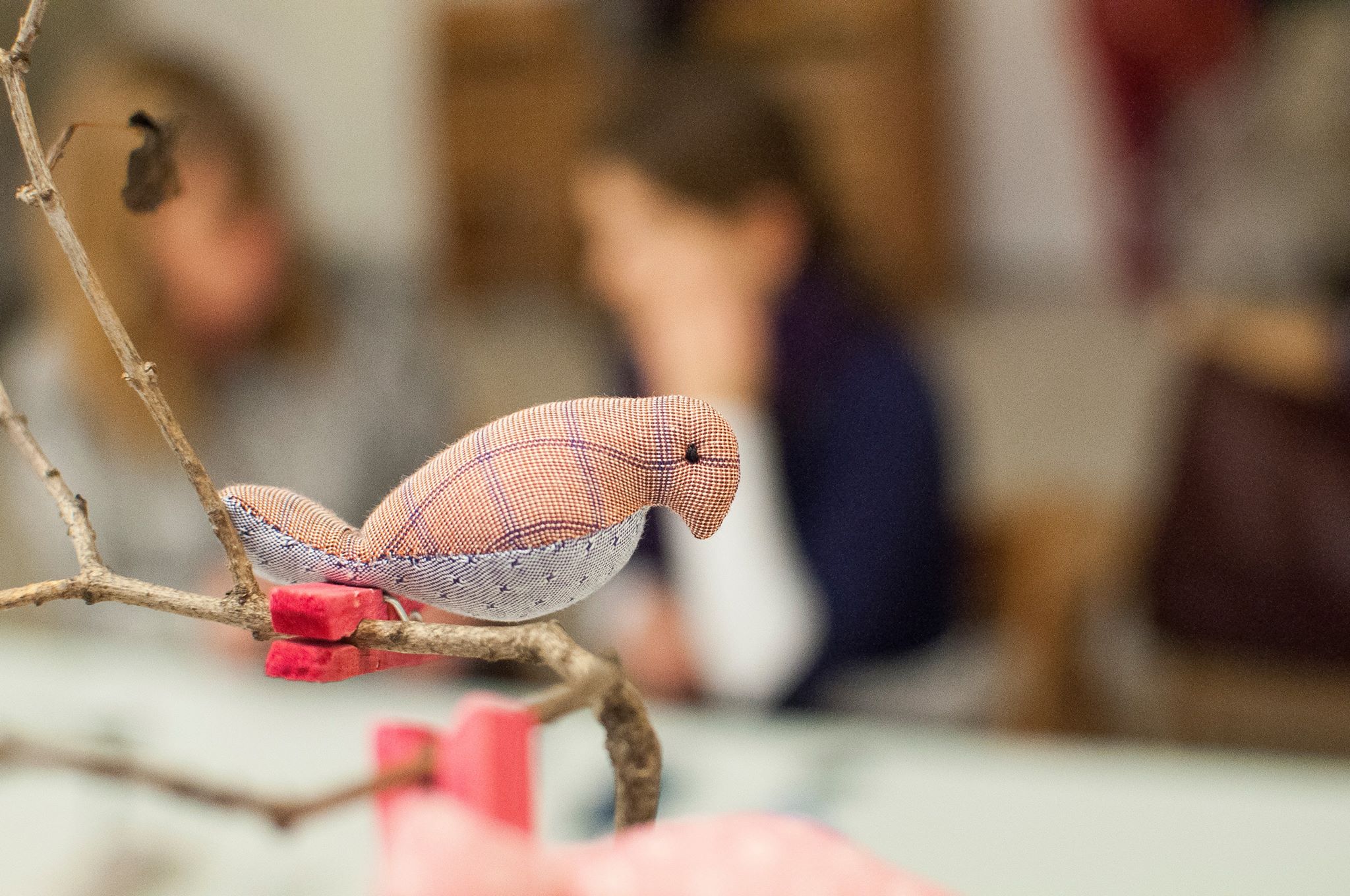 na zdjęciu wyostrzone zbliżenie na uszytego ręcznie ptaszka w różowych barwach; ozdoba jest zamocowana na gałązce; w tle uczestnicy zajęć pracujące nad nowym rękodziełem