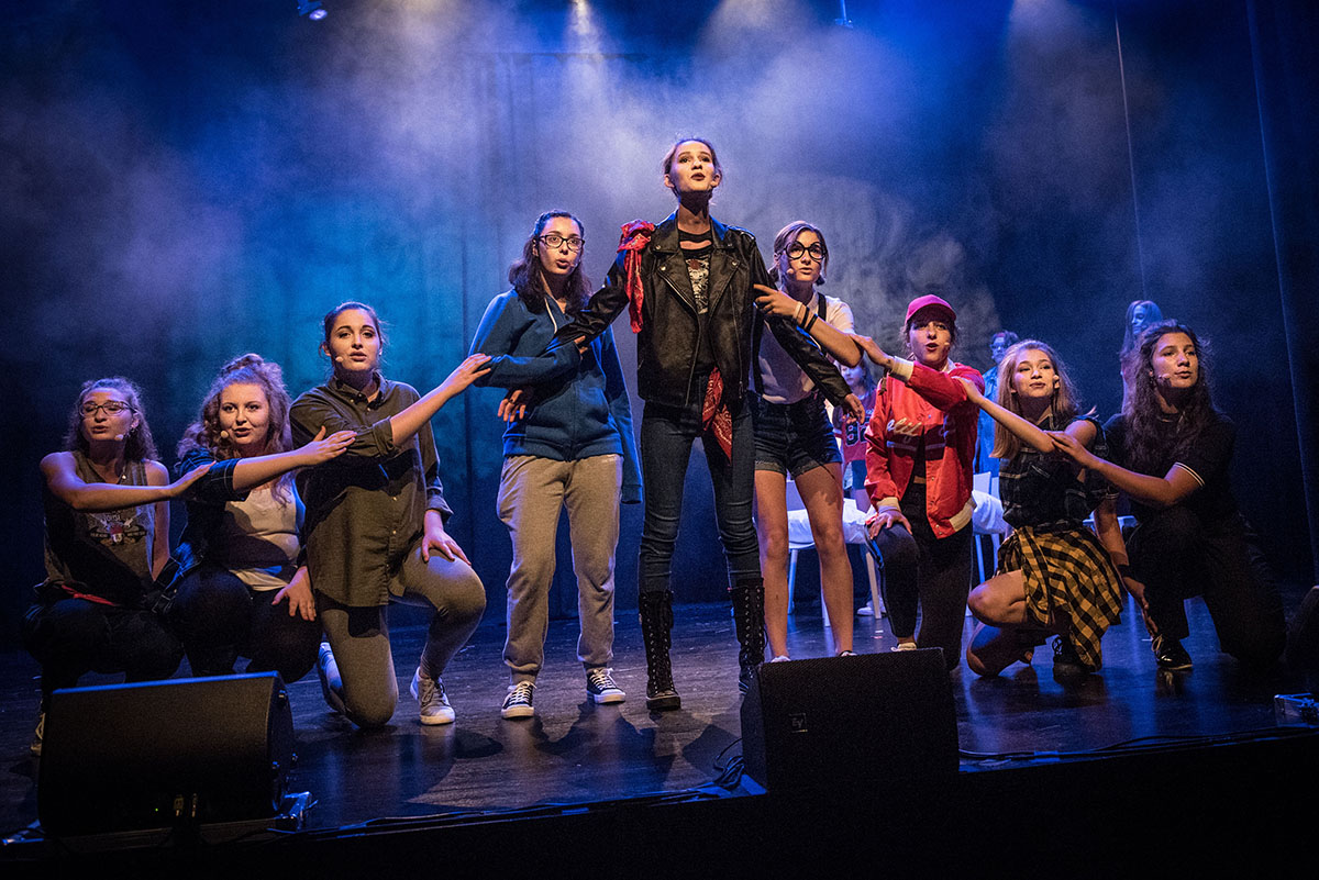 Zdjęcie promocyjne pracowni. Fot. Marta Ankiersztajn Spektakl "Hejt Made"  / Na zdjęciu widoczna jest grupa młodych dziewcząt na scenie teatralnej. Dziewczyny stoją w linii na skraju sceny.