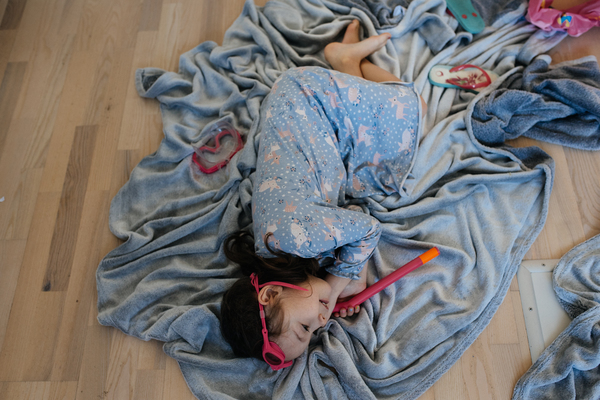 Na zdjęciu dziewczynka leżąca na podłodze na dużym szarym kocu. Dziewczynka ma na głowie maskę do nurkowania, a w dłoni trzyma rurkę do nurkowania.