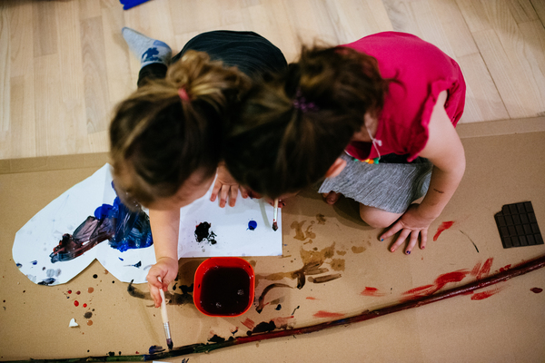 Na zdjęciu widać dwoje dzieci malujących karton farbami. Koło nich leży długi patyk. 