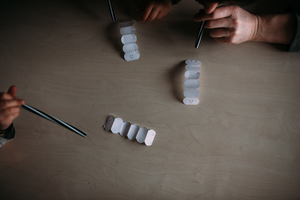 Na zdjęciu widać trzy papierowe harmonijki na kształt dżdżownic oraz trzy dłonie ze słomkami, które dmuchają w ich kierunku. 