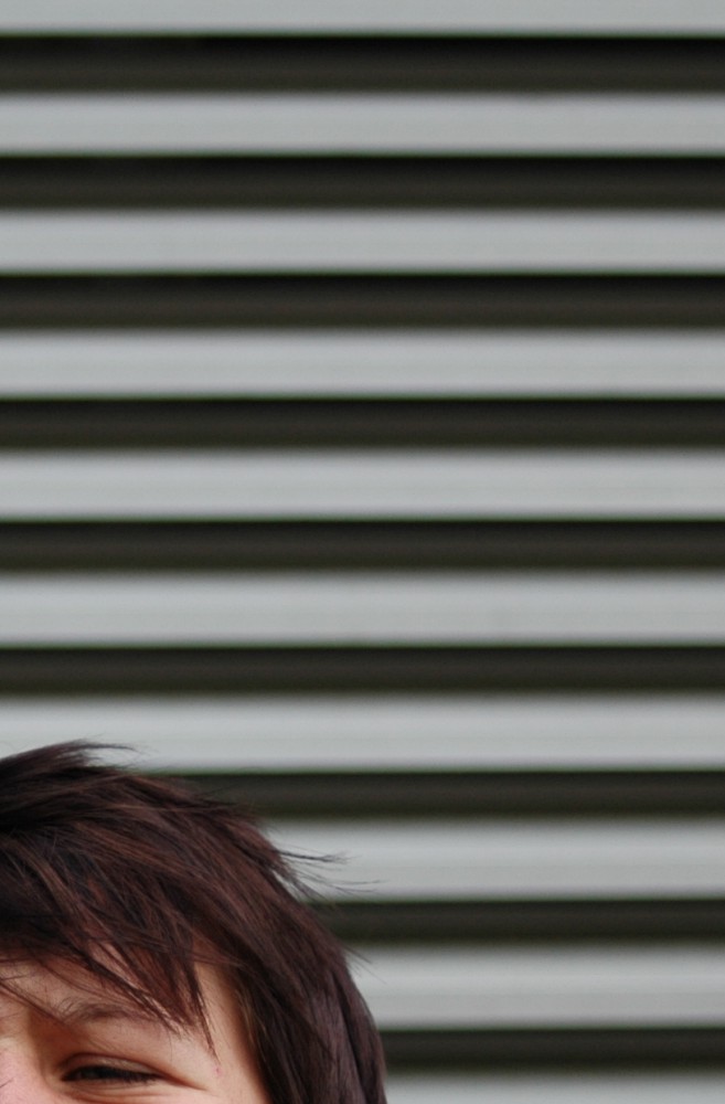 Zdjęcie zdominowane przez poziome szare pasy ułożone naprzemiennie w poziomie. W lewym dolnym rogu widać fragment twarzy młodej osoby niemożliwej do indetyfikacji. Ćwierć twarzy znajduje się w lewym dolnym rogu, włosy brązowe, krótkie, opadają na czoło, oczy przymarszczone. Foto: Joanna Kinowska