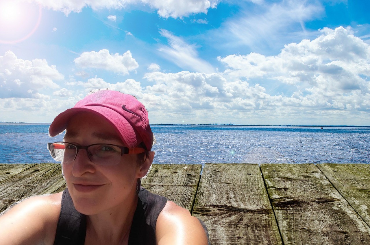 Na zdjęciu doklejona autroportret (selfie) do widoku na morze lub ocean opisany na poprzedniej fotografii. Bohaterka znajduje się w lewej części zdjęcia, ma ręce wyciągnięte do wykonania pamiątki fotograficznej. Na sobie ma różową czapkę z daszkiem i okulary korekcyjne.