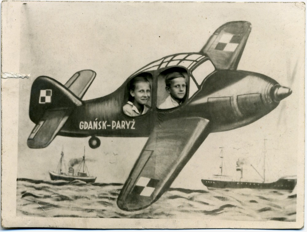 Dwóch chłopców (nastolatków) pozuje do zdjęcia z namalowanym samolotem z napisem "Gdańsk-Paryż". Dzieci pozują ustawione za dużym, wymalowanej płachcie z wyciętymi dziurami symulującymi szyby samolotu. Fotografia w odcieniach szarości i sepii.