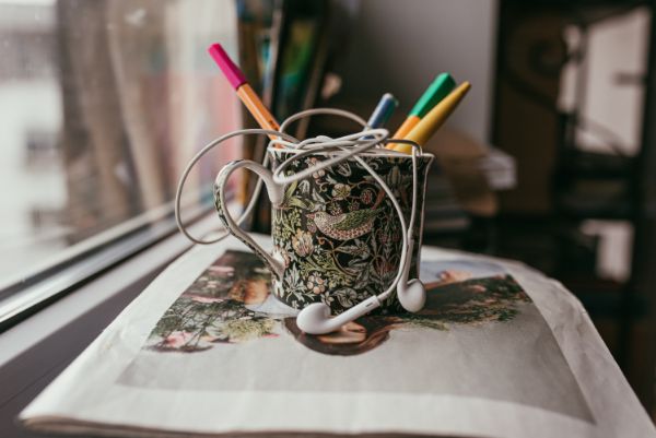 Na parapecie, na stosie gazet stoi kubek po kawie, zawiera: dwa kolorowe cienkopisy, dwa długopisy oraz białe słuchawki douszne. Fot. Agnieszka Mocarska