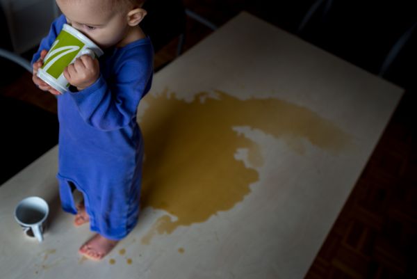 Zdjęcie zrobione od góry przedstawia 1,5-roczne dziecko w rozpiętym ubranku stojące w wielkiej plamie kawy na stole i dopijające resztki z kubeczka. Fot. Agnieszka Mocarska