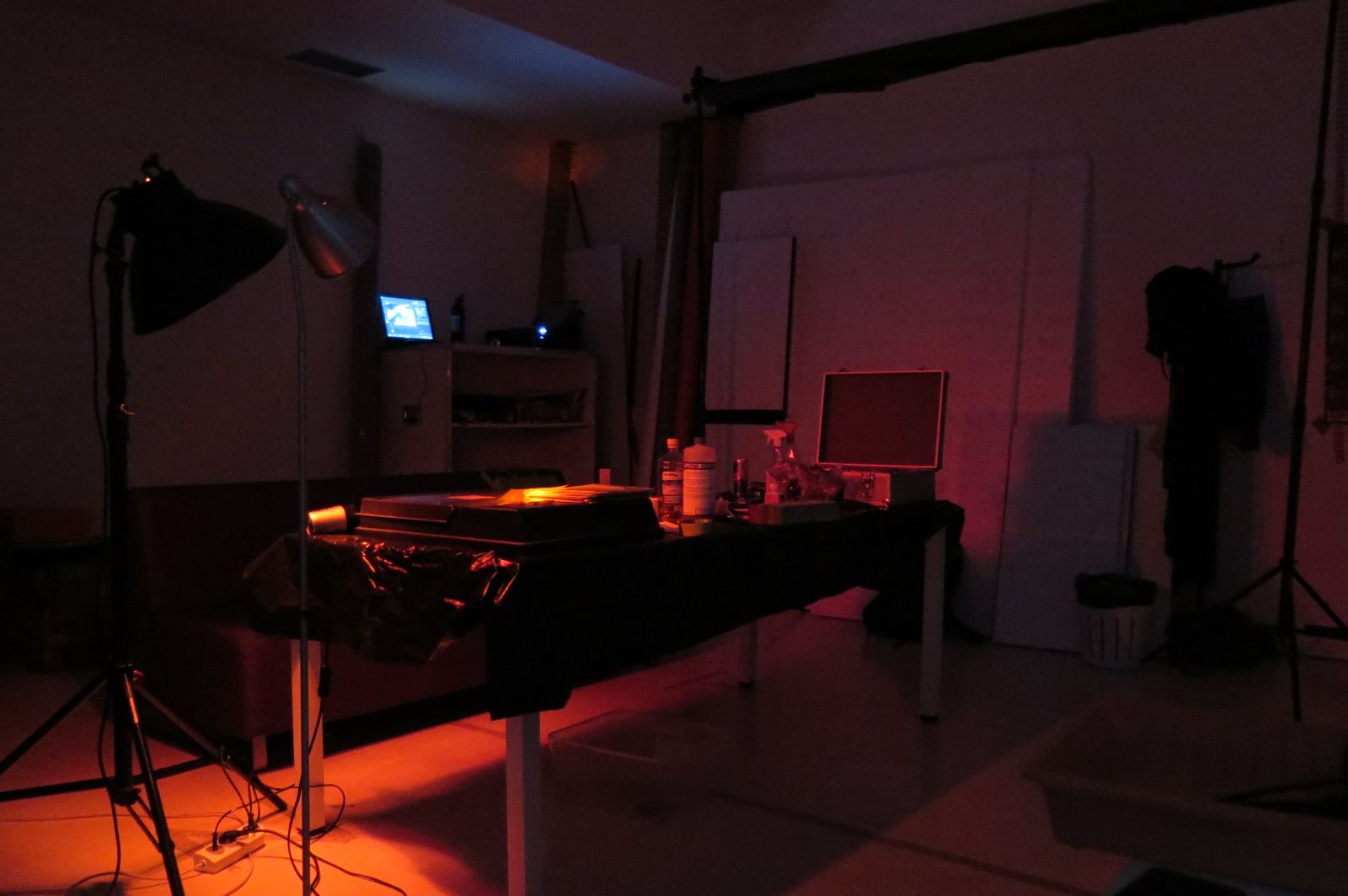 Zdjęcie promocyjne pracowni.  Na zdjęciu studio fotograficzne z przyciemnionym, czerwonym światłem. Na środku studia znajduje się stół oraz kilka lamp studyjnych.
