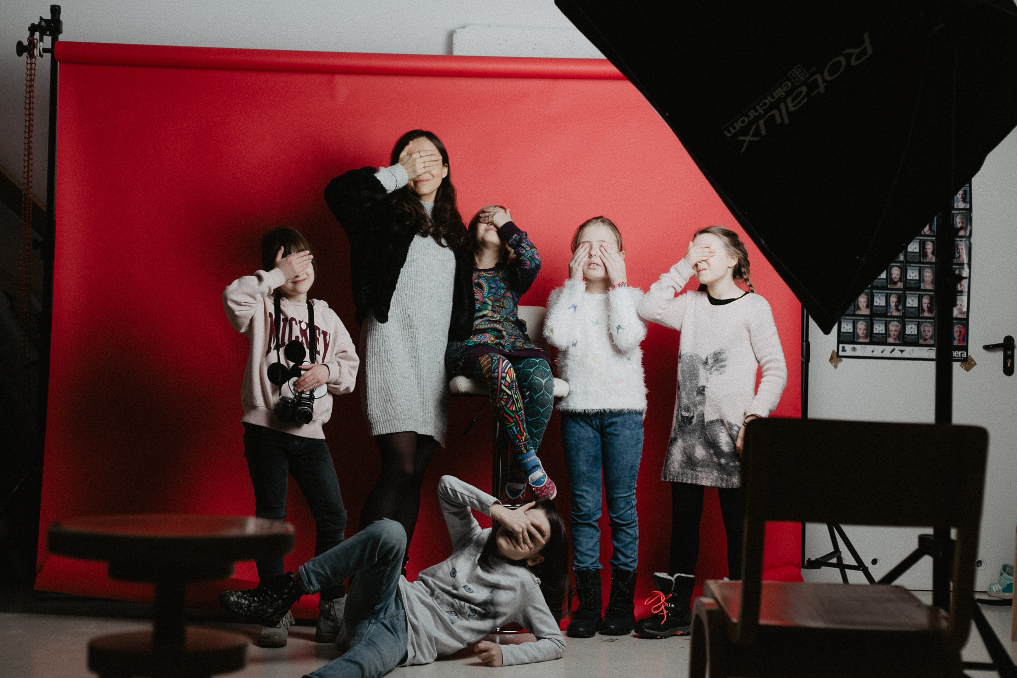 Fotografia wykonana w trakcie warsztatów Na zdjęciu Marta Rybicka - instruktorka zajęć fotograficznych - wraz z piątką dzieci. Wszyscy znajdują się w studiu foto, mają zasłonięte twarze, co uniemożliwia ich identyfikacje. W tle jest rozłożona czerwone tło do sesji fotograficznych, po prawej stronie fragment lampy studyjnej.
