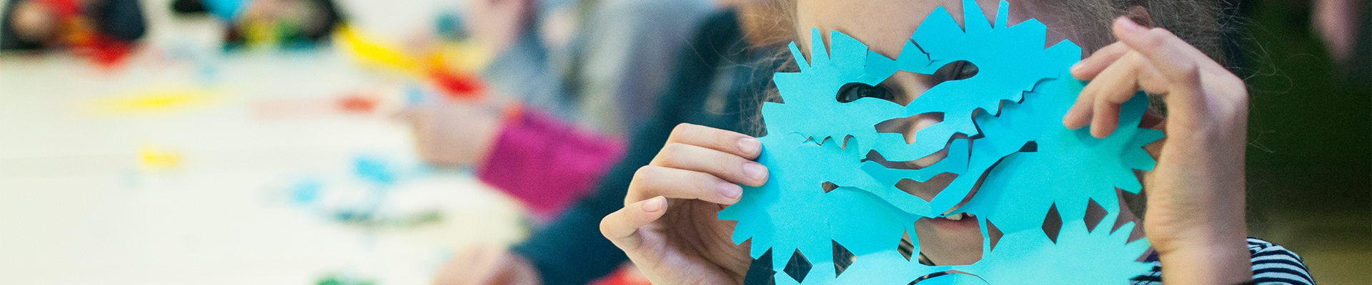 Na zdjęciu dziewczyna zasłania twarz swoją niebieską wycinanką. W tle po lewej stronie widok na pracownię plastyczną.