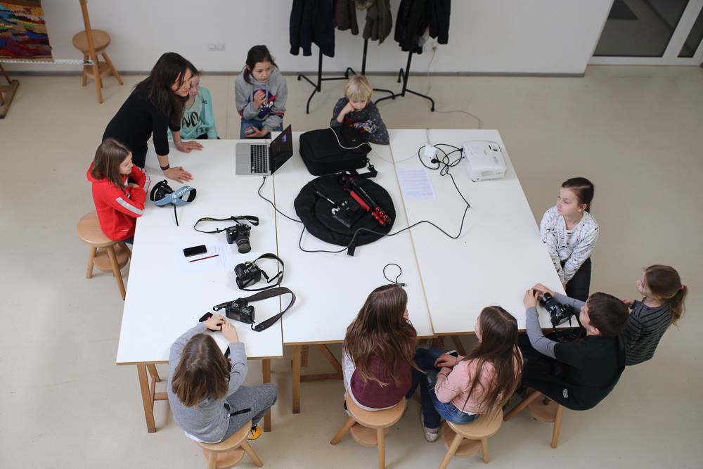 Ujęcie z góry na wielki stół, przy nim grupa dzieci i instruktorka podczas zajęć fotograficznych. Na stole leży sprzęt fotograficzny. Fot. Łukasz Wielgo