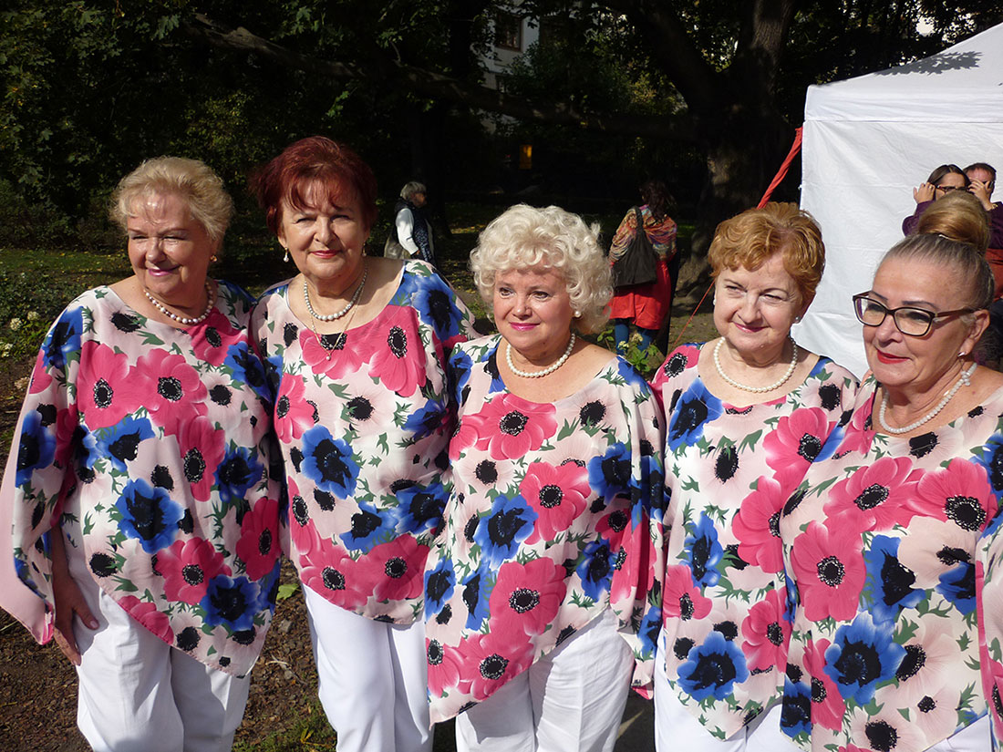 Występy w ogrodzie Saskim. Na zdjęciu 5 wokalistek zespołu Służewianki pozuje na tle pikniku. Ubrane w białe spodnie oraz kolorowe bluzki z wzorami kwiatów. 