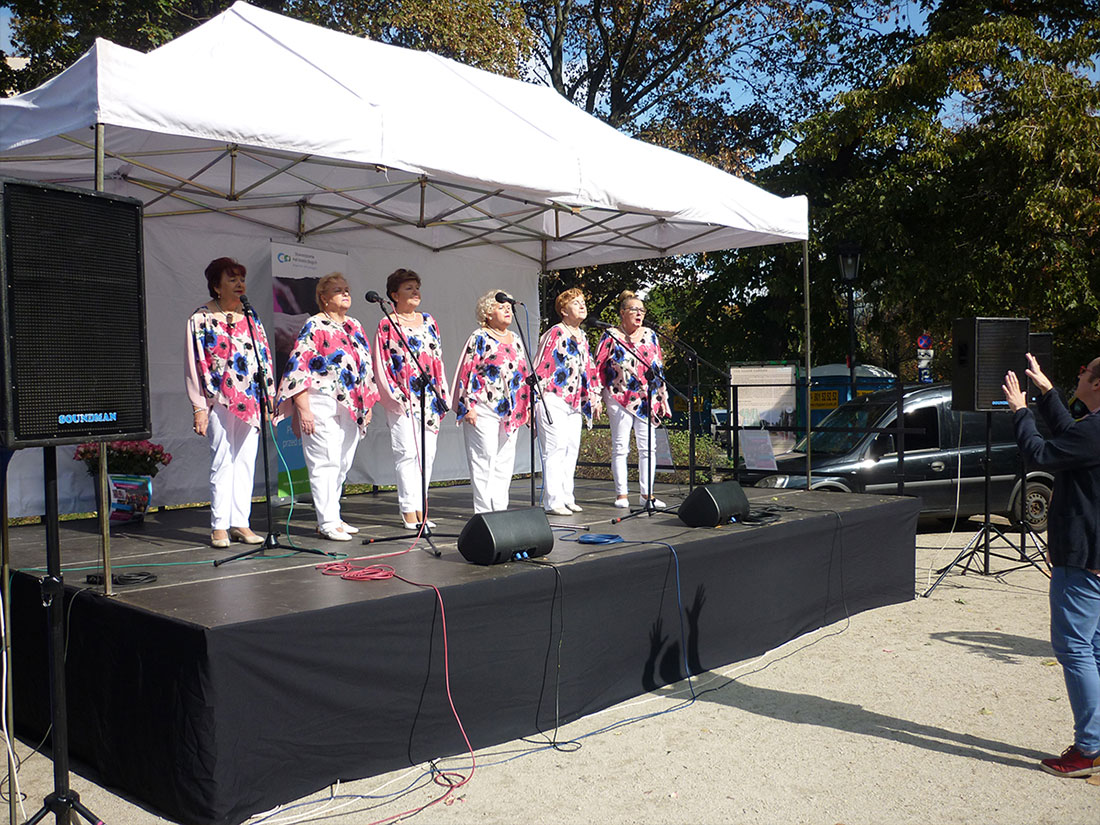 Zdjęcie z występów w ogrodzie Saskim. 6 wokalistek zespołu Służewianki na scenie. Ubrane w białe spodnie oraz kolorowe bluzki z wzorami kwiatów. Przed sceną prowadzący zespół gestami udziela rad.