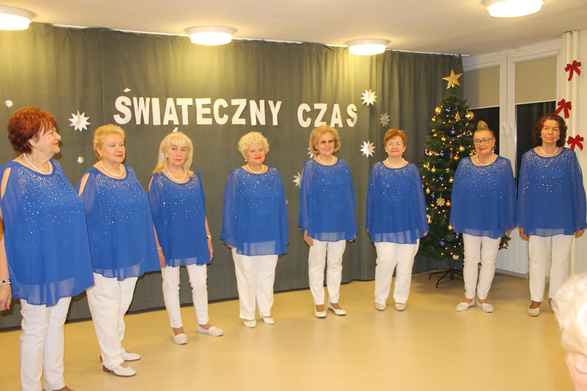 Na zdjęciu grupa wokalistek zespołu Służewianki stoi w półkolu. Ubrane w białe spodnie oraz niebieskie, luźne bluzki.