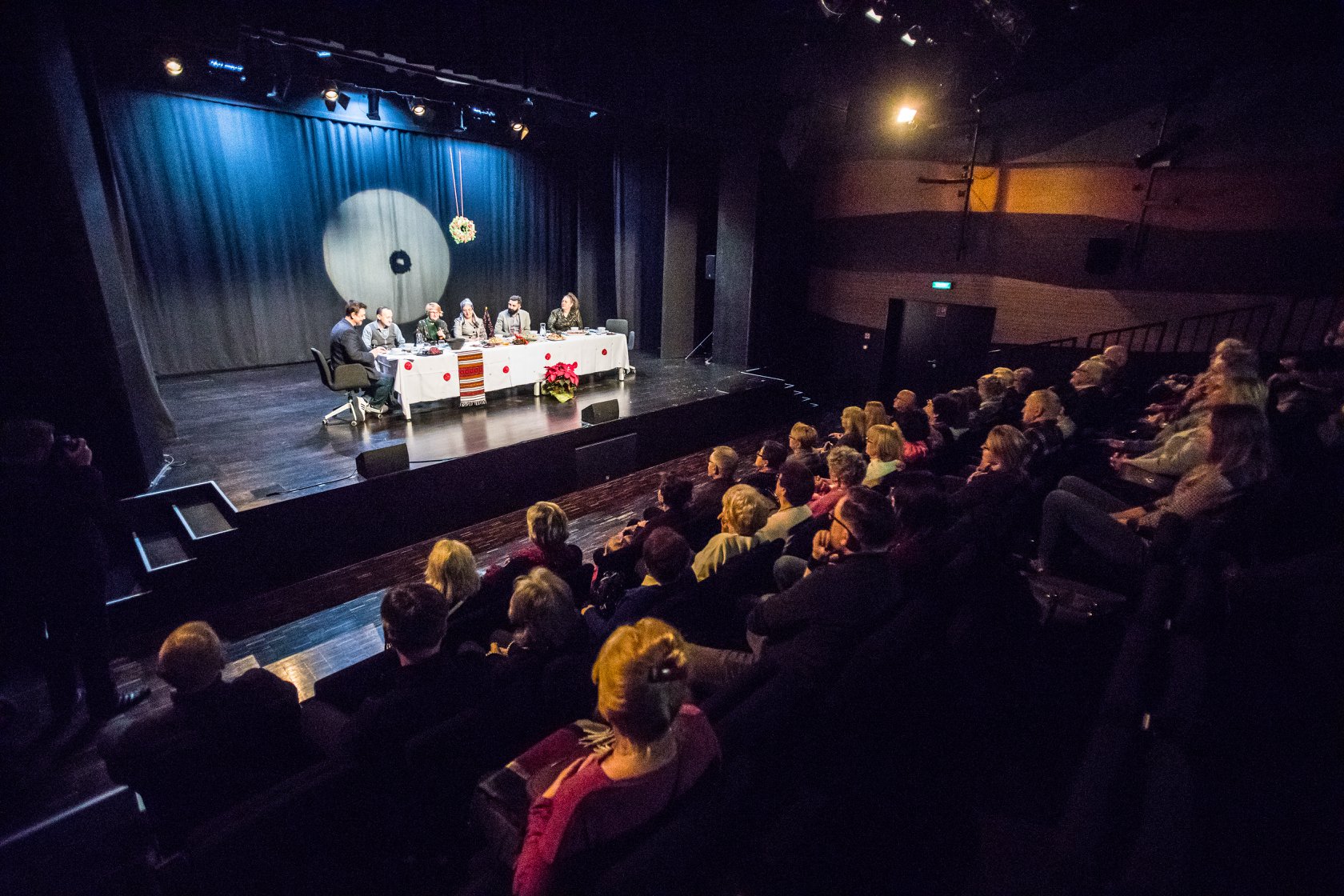 Na pierwszym planie słuchająca widownia, w tle scena SDK, na niej oświtlony biały stół, wokół którego usiedli goście wigilii wielokulturowej