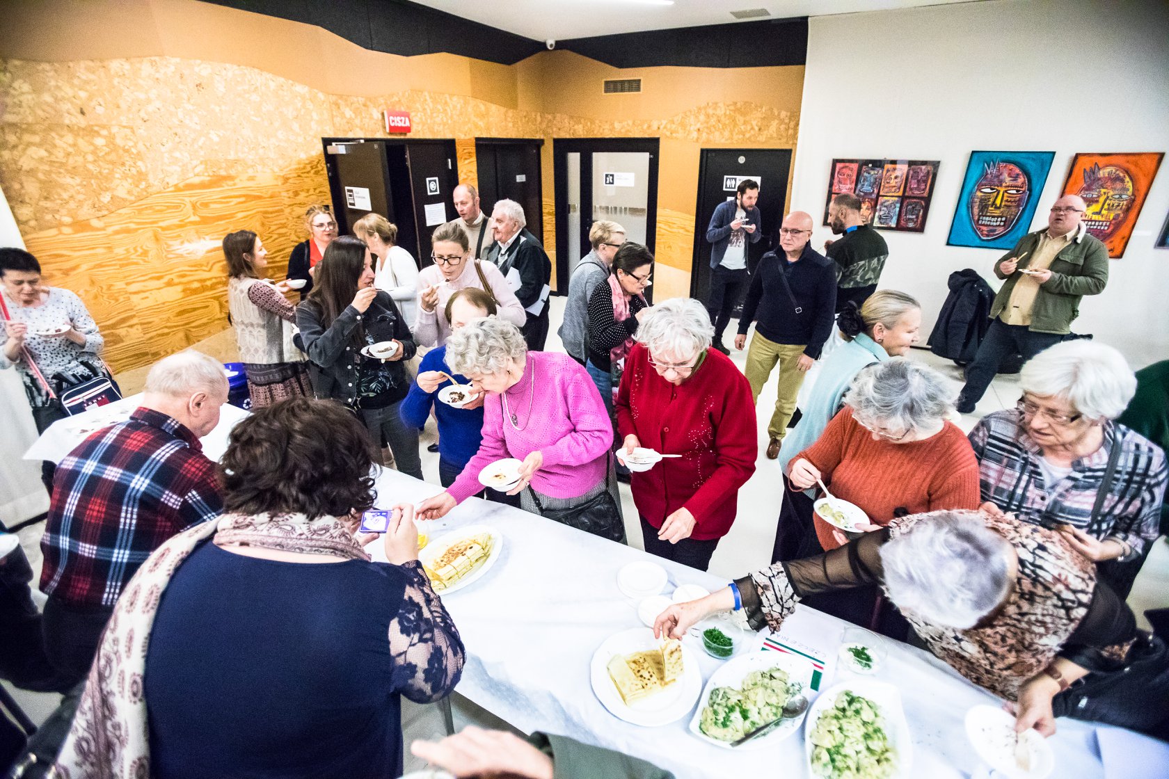 Na zdjęciu widoczny poczęstunek z prezentowanych dań wigilijnych. Na poerwszym planie biały stół z potrawami, w tle uczestnicy spotkania jędzący potrawy 