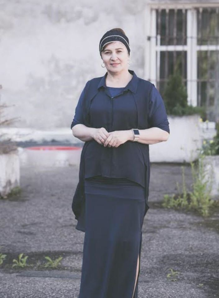 Na zdjęciu portret ogólny kobiety ubranej na granatowo, z zasłoniętymi włosami opaską, w tle szara kamienica