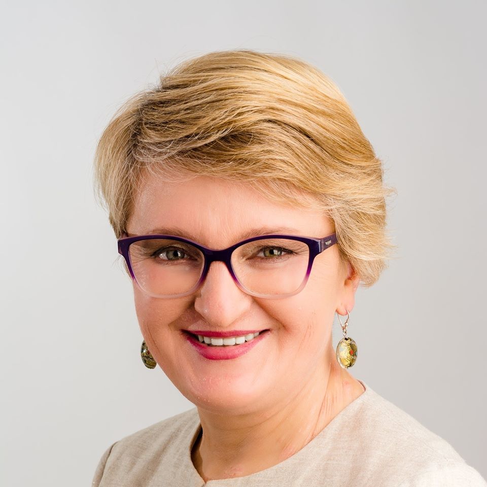 Na zdjęciu krótko ostrzyżona blondynka w okularach, w kolczykach i pomalowanymi na różowo ustami