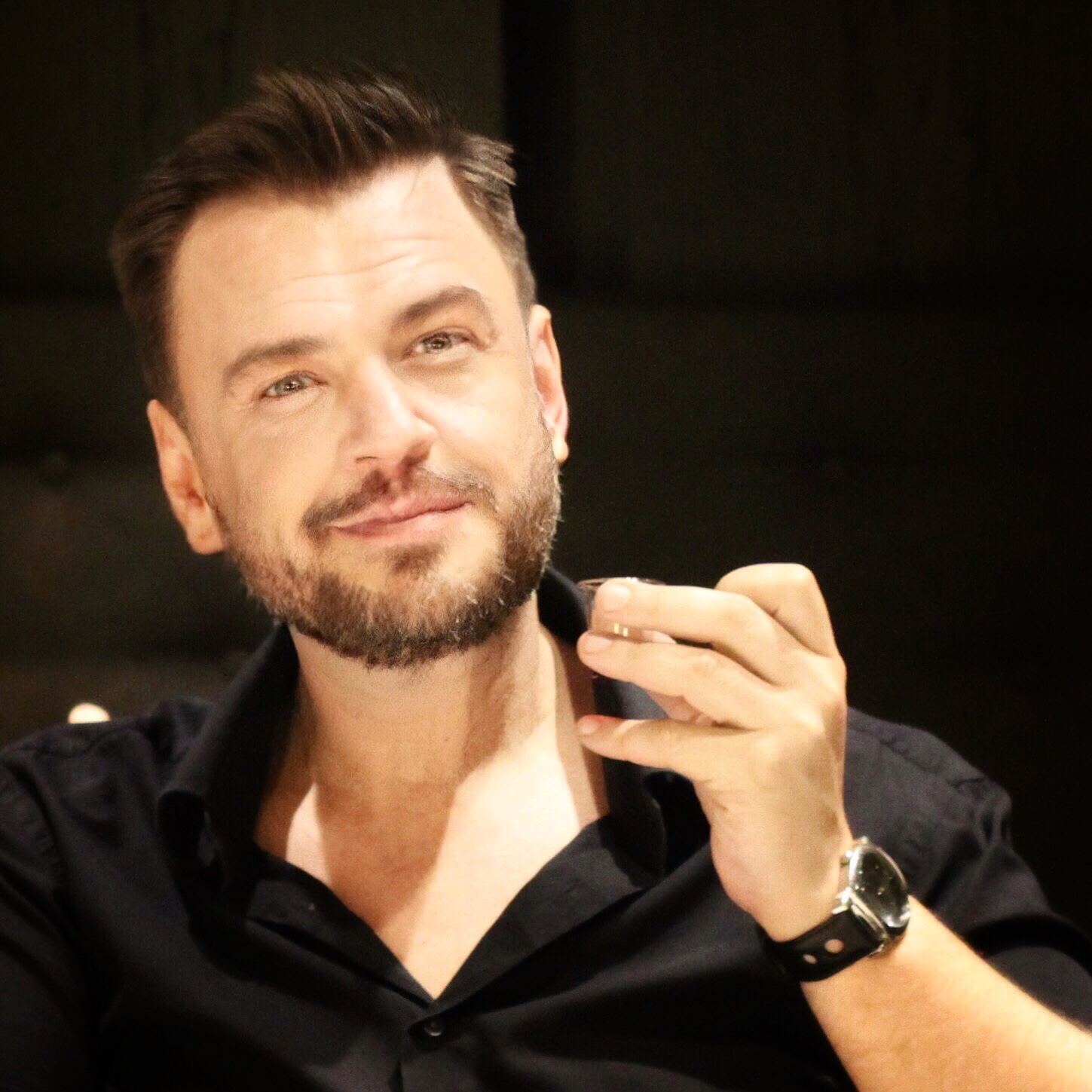 portret mężczyzny w czarnej koszuli, z lewą ręką uniesioną na wysokości brody.