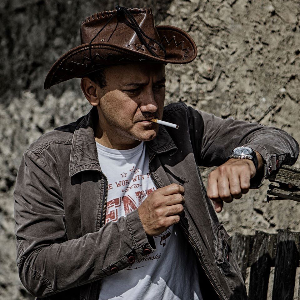 Na zdjęciu mężczyzna w kapeluszu kowbojskim, z papierosem w ustach. W tle szara, kamienna sceneria
