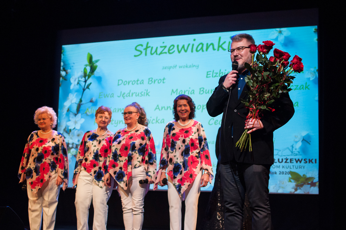 Na zdjęciu Burmistrz Rafał Miastowski oraz 4 członkinie zepołu wokalnego, które zaraz otrzymają róże po występie. Burmisrz jest ubrany na ciemno i trzyma mikrofon, wokalistki są ubrane w białe spodnie i kwieciste bluzki. W tle grafika z wymienionymi nazwiskami wokalistek, wymienione w artykule.