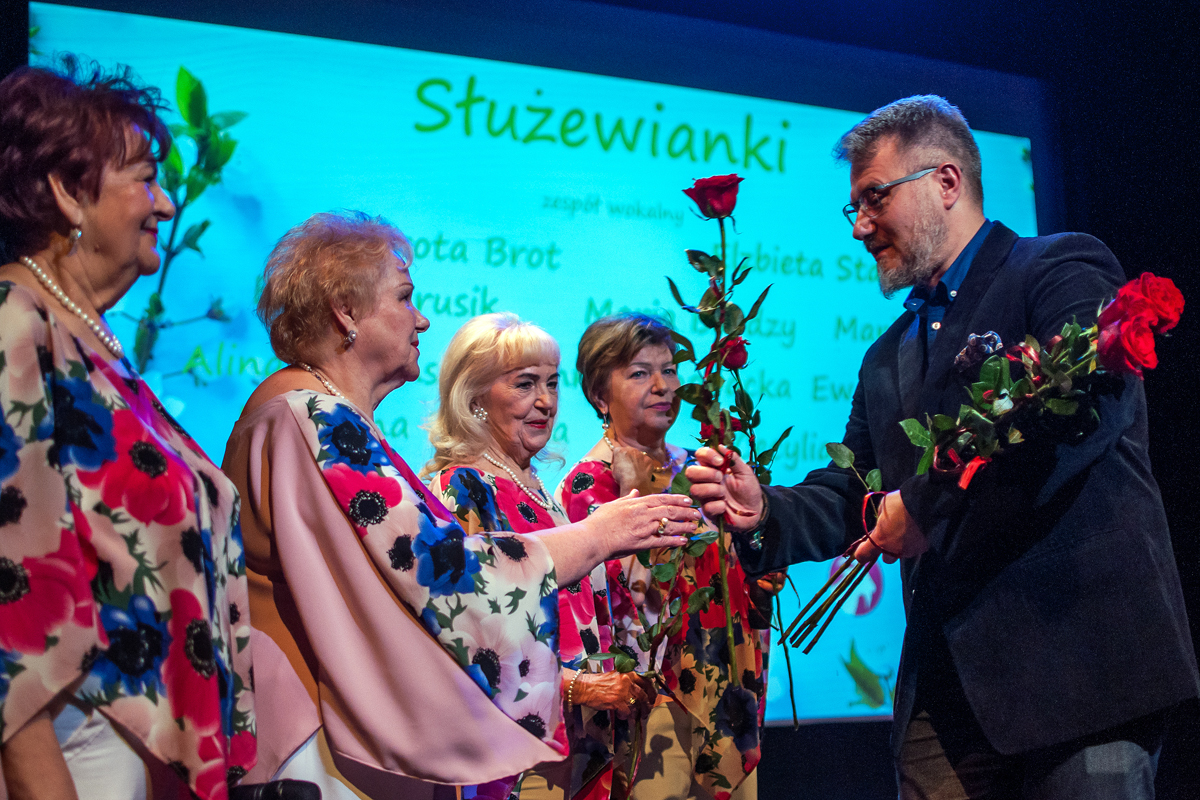 Na zdjęciu 4 członkinie zepołu wokalnego otrzymują róże od Burmistrza Rafała Miastowskiego. Burmisrz jest ubrany na ciemnon, wokalistki są ubrane w kwieciste bluzki. W tle grafika z wymienionymi nazwiskami wokalistek, wymienione w artykule.