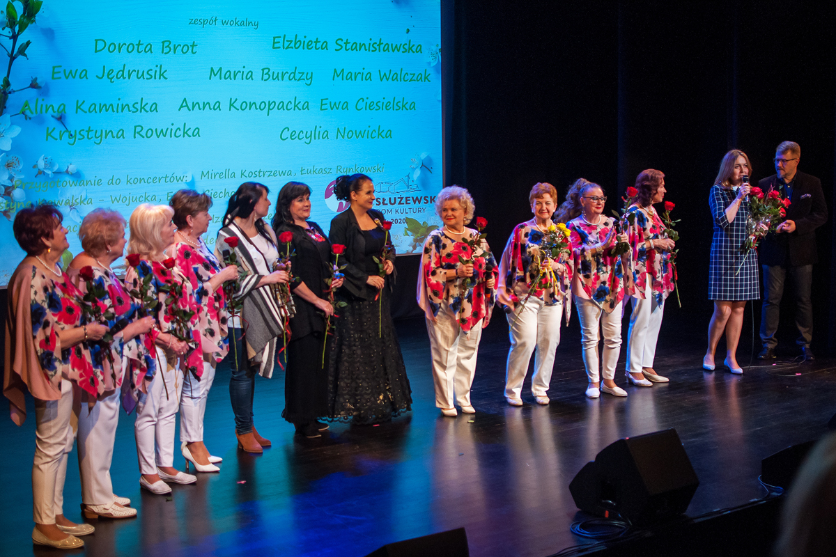 Na zdjęciu wszystkie członkinie zepołu wokalnego weraz z instruktorami i Burmistrzem Mokotowa. , które zaraz otrzymają róże po występie. Jedna z osób przemawia do widownii. W tle grafika z wymienionymi nazwiskami wokalistek, wymienione w artykule.
