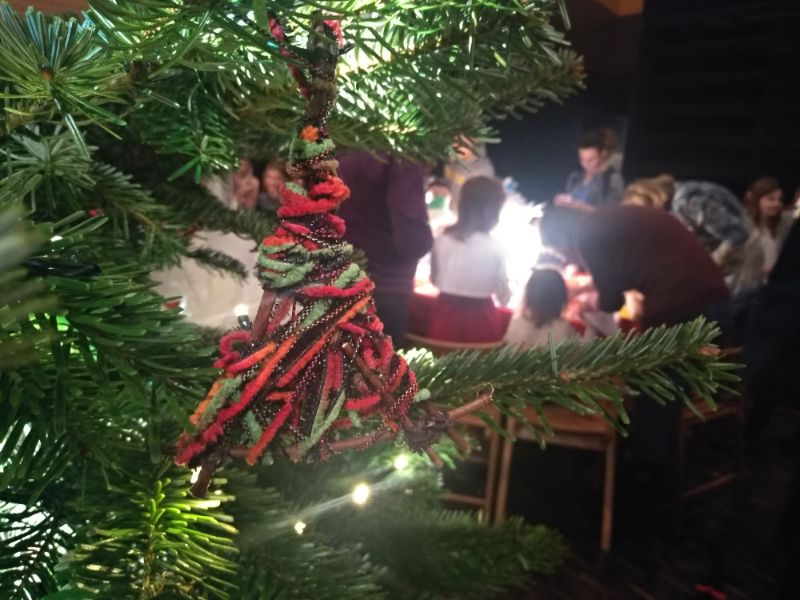 Zdjęcie przedstawia przygotowaną przez jednego z uczestników warsztatu plastycznego ręcznie wykonaną ozdobę choinkową. W tle choinka i pracująca grupa warsztatowa.