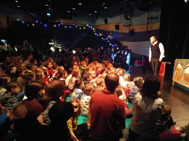 Na zdjęciu tłum dzieci (nie do zidentyfikowania) oglądające przedstawienie mikołajkowe. W tle przyciemniona sala widowiskowa.