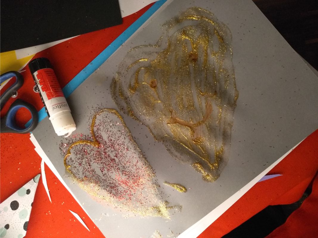 Zdjęcie przedstawia pracę plastyczną z dwoma namalowanymi sercami. W tle czerwony obrus i przybory papiernicze