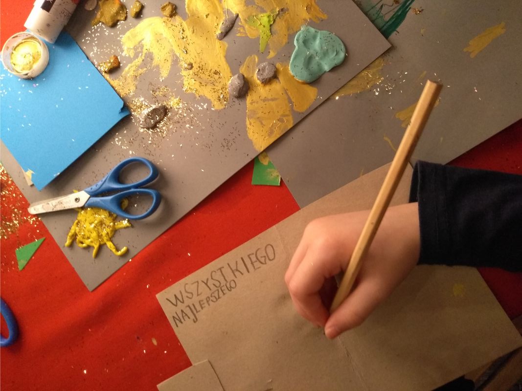 Zdjęcie przedstawia przygotowywaną pracę plastyczną z napisem "wszystkiego najlepszego". W tle czerwony obrus i inne prace plastyczne